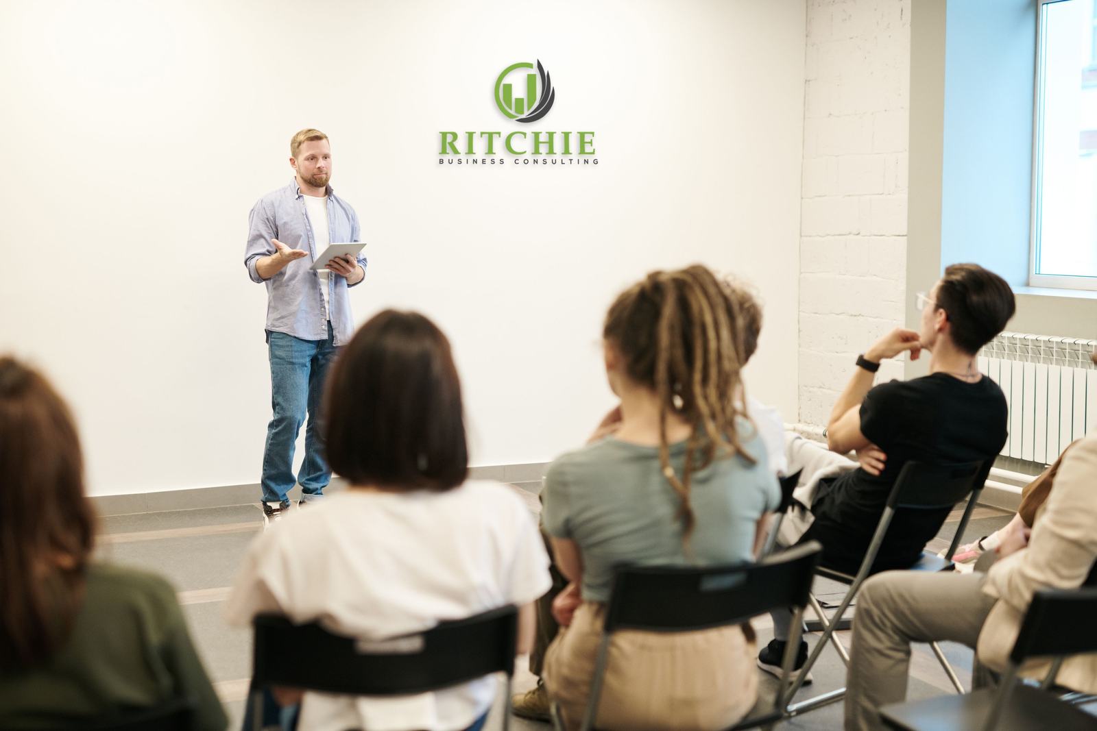 A man in a button-down shirt leading a small meeting, with the Ritchie Business Consulting mark on the wall.