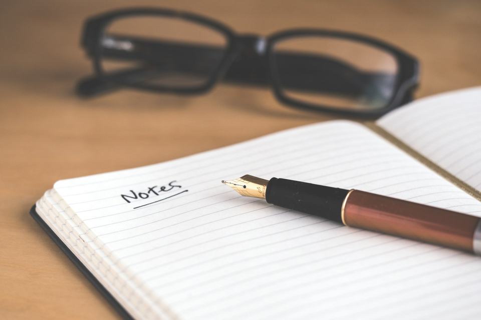 An open notebook labeled Notes, a fountain pen, and eyeglasses on a wooden desk.